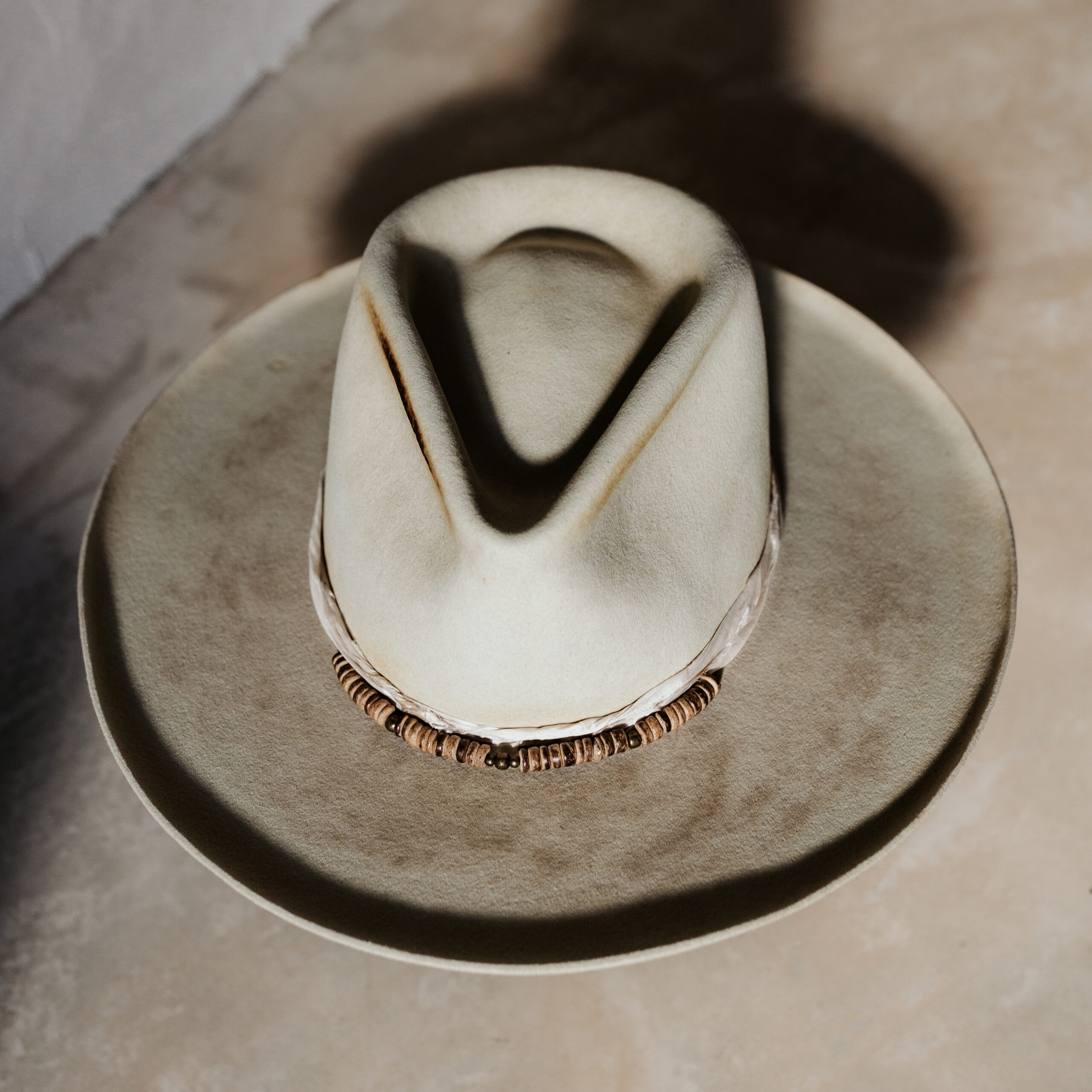 A beautifully handcrafted Crown of the Vagabond hat by the hatters Ruth and Chris on Mallorca with traditional tools in the color Vanilla shaped in Gold Digger with a naturally distressed design consisting of a silk cord tied in the back as well as wood pearls threaded in the front and burn marks on the crown with a curled brim - branded with the serial number 19 made during our image video.