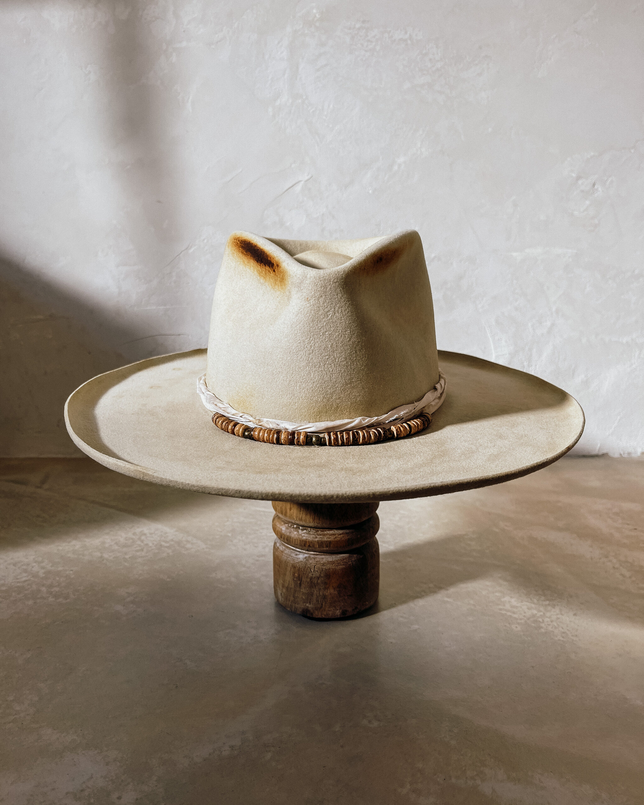 A beautifully handcrafted Crown of the Vagabond hat by the hatters Ruth and Chris on Mallorca with traditional tools in the color Vanilla shaped in Gold Digger with a naturally distressed design consisting of a silk cord tied in the back as well as wood pearls threaded in the front and burn marks on the crown with a curled brim - branded with the serial number 19 made during our image video.