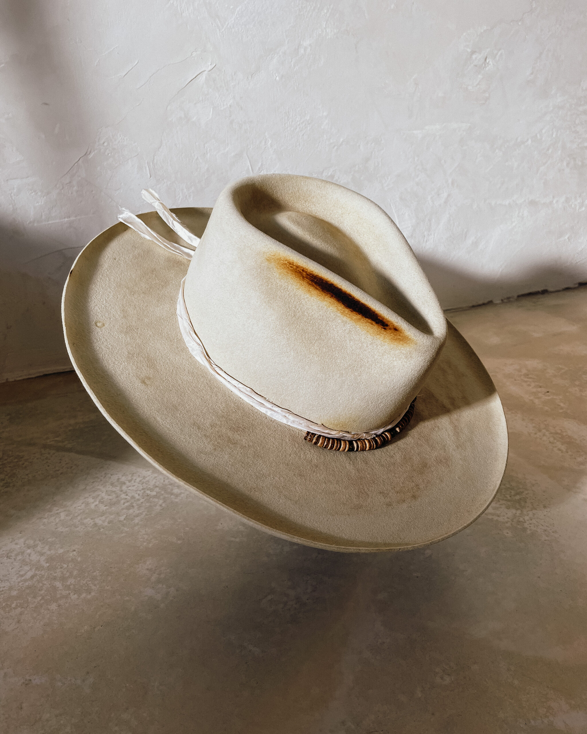 A beautifully handcrafted Crown of the Vagabond hat by the hatters Ruth and Chris on Mallorca with traditional tools in the color Vanilla shaped in Gold Digger with a naturally distressed design consisting of a silk cord tied in the back as well as wood pearls threaded in the front and burn marks on the crown with a curled brim - branded with the serial number 19 made during our image video.