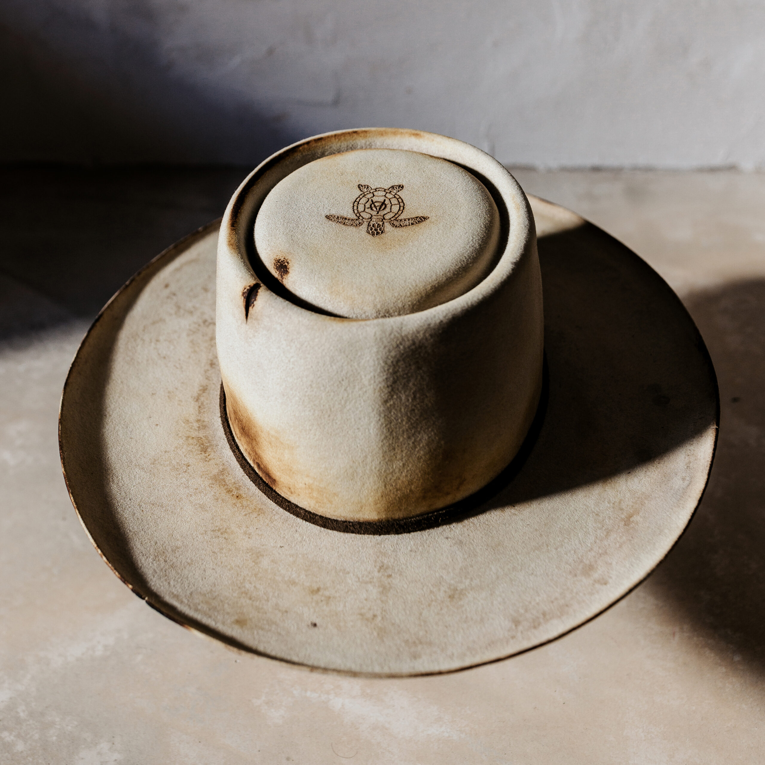 A beautifully handcrafted Crown of the Vagabond hat by the hatters Ruth and Chris on Mallorca with traditional tools in the color Vanilla shaped in Oasis with a distressed used look consisting of burn marks as well as cutouts tied on the crown with a rough leather string fixed with a bronze pearl in the back and a turtle engraved on the top of the crown and a curled brim - branded with the serial number 22.