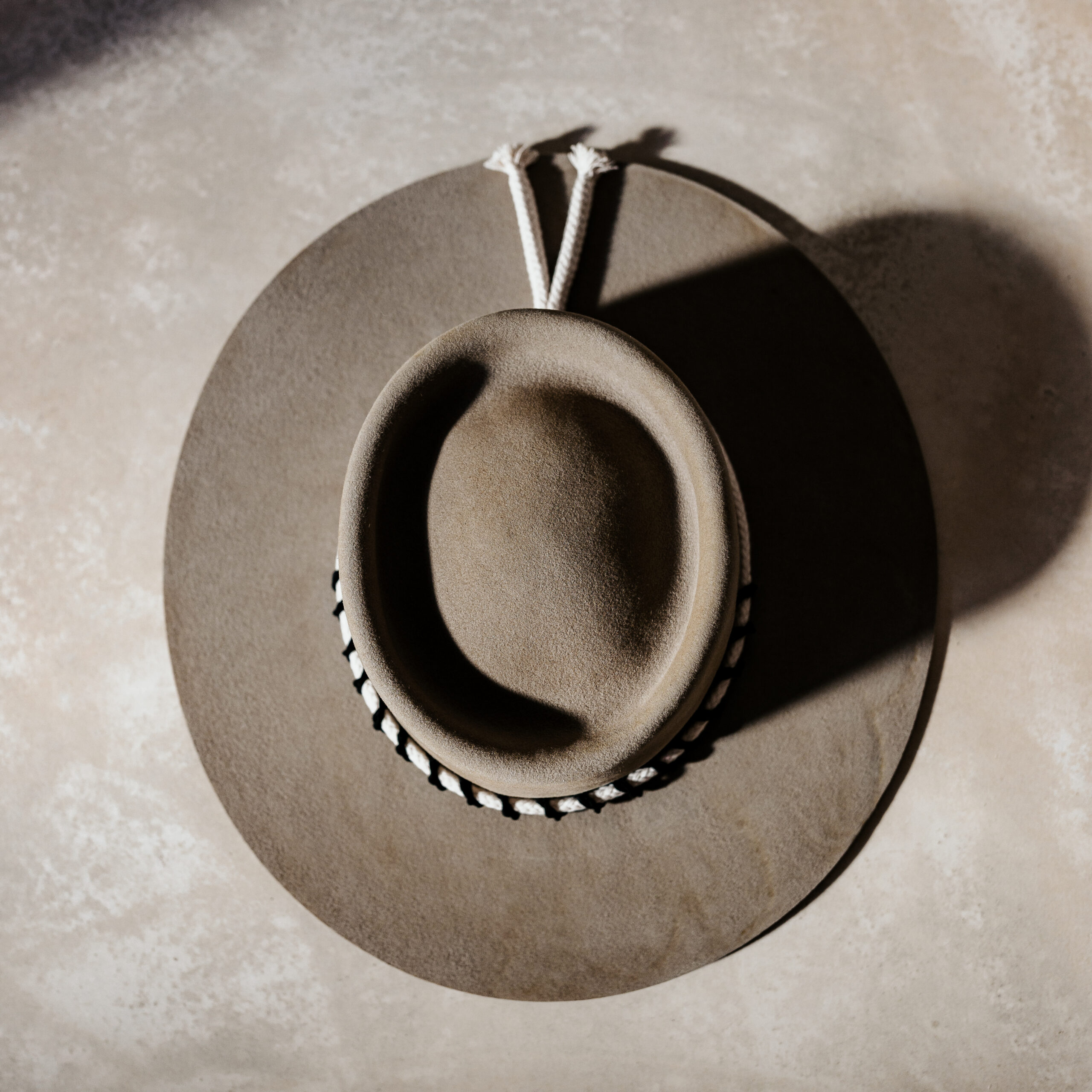 Crown of the Vagabond customised handcrafted hat by Ruth and Chris with traditional tools made in Mallorca, Porto Cristo. Oasis shaped in Prairie Dust with a flat brim in a simple greek style with a white robe surrounded by a black corded leather string which is also used as the chinstrap fixed with a wooden pearl.