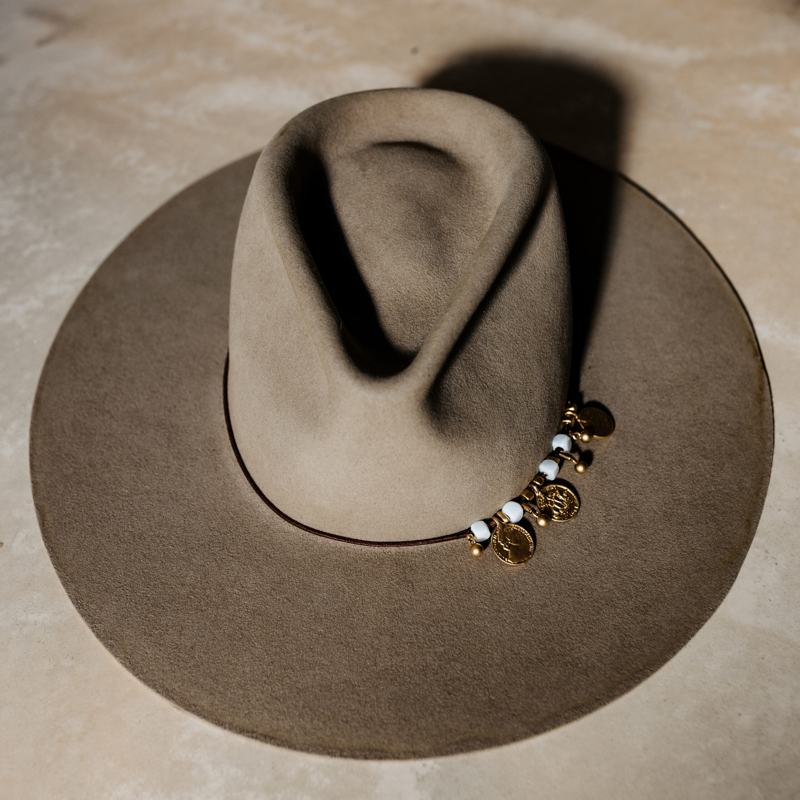 A beautifully handcrafted Crown of the Vagabond hat by the hatters Ruth and Chris on Mallorca with traditional tools in the color Prairie Dust and shaped in Gold Digger with an opulence look inspired by the antique and old ages consisting of a brown leather cord threading golden coins as well as golden other small golden elements and white formed stones with a flat brim - branded with the serial number 8.