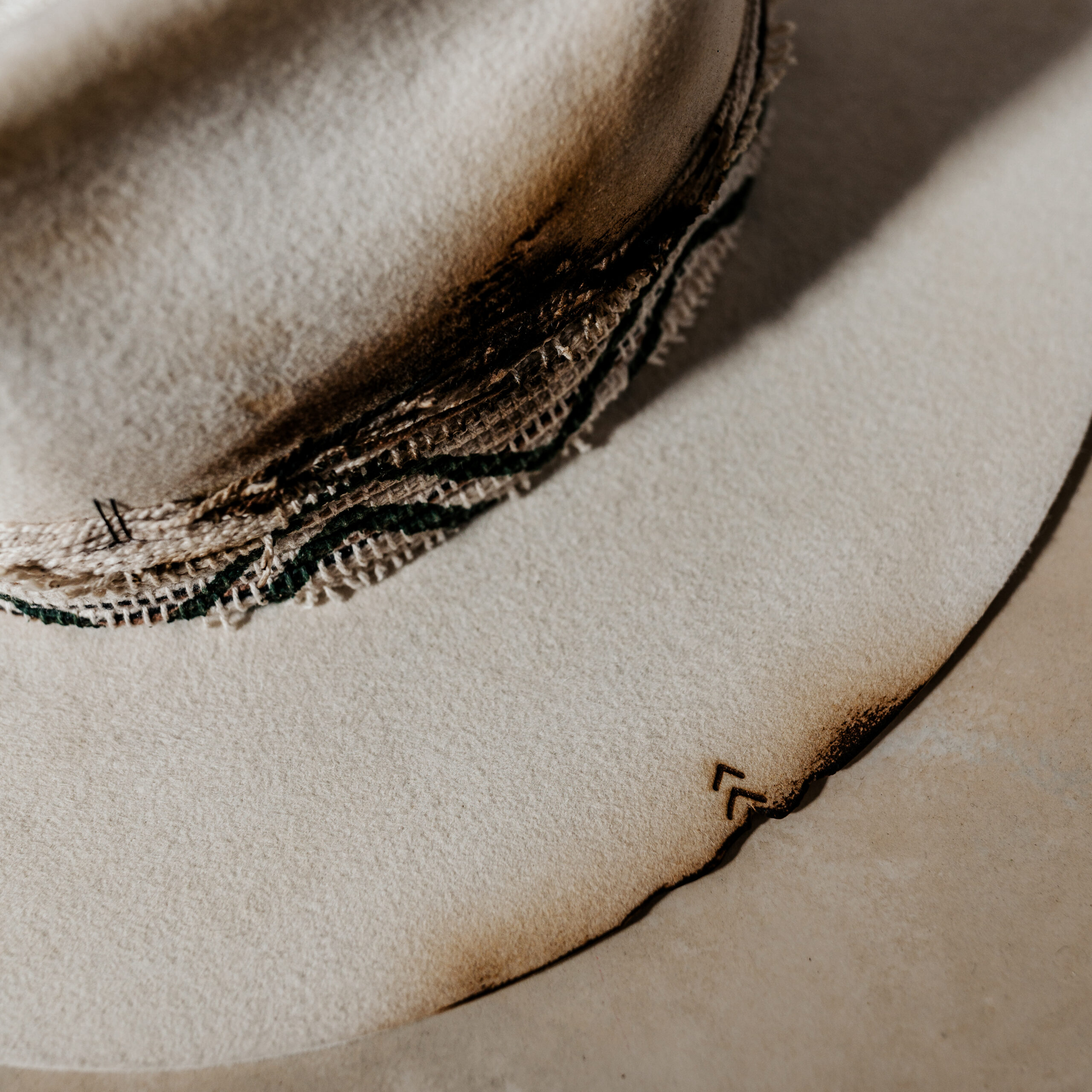 A beautifully handcrafted Crown of the Vagabond hat by the hatters Ruth and Chris on Mallorca with traditional tools in the color Snow shaped in Stargazer with a rough adventurer look mostly distressed consisting of a woven thick headband with stitches on crown and band as well as custom laser branding on crown and brim near the cutouts. The flat brim has cutouts as well as burn marks all over and the sweatband wears the number 28.